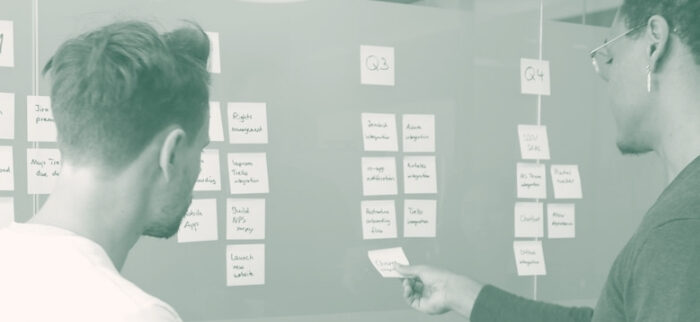 Zwei Kollegen stehen vor einer Pinnwand mit Post-its und delegieren Aufgaben.