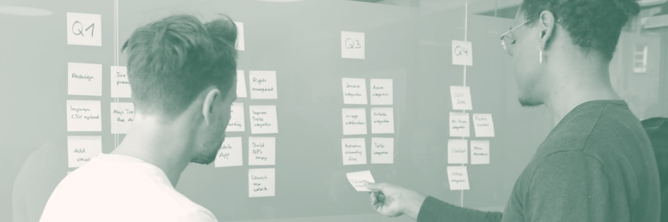 Zwei Kollegen stehen vor einer Pinnwand mit Post-its und delegieren Aufgaben.