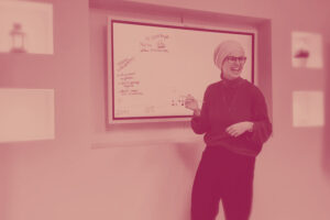 Eine Frau mit Brille und Kopftuch steht laechelnd vor einem Whiteboard. Sie haelt einen Stift in der Hand und scheint etwas zu erklaeren.