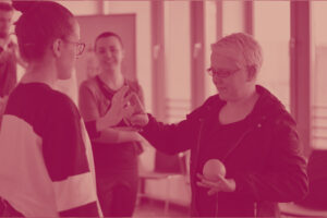 In einem Teamtraining stehen drei Frauen zusammen. Eine Teilnehmerin wird gefuehrt und haelt eine Orange in einer Hand, waehrend sie mit der anderen die Hand der Trainerin beruehrt.